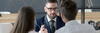 a man on the other side of the table wearing a suit and tie talking to a woman and a man